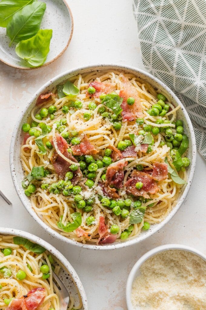 Low bowl filled with pasta with bacon, peas, and a light emulsified olive oil-based sauce, topped with Parmesan, fresh basil, and black pepper.