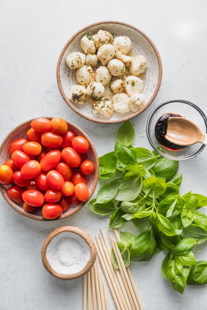 Overhead image of cherry-sized mozzarella balls, cherry tomatoes, lots of fresh basil, skewers, sea salt, and balsamic glaze.