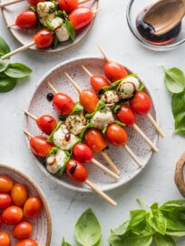 Countertop set with plates of Caprese skewers, extra tomatoes, fresh basil, and balsamic glaze.