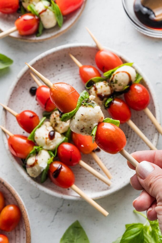 Close up of a hand lifting a Caprese skewer from a small appetizer plate.