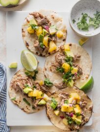 White platter holding four carnitas tacos with pineapple jalapeno salsa and diced avocado.