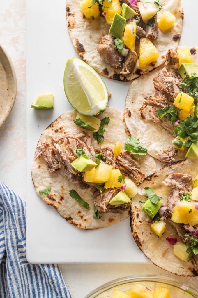 Instant Pot carnitas tacos with pineapple salsa and diced avocado, assembled and served with lime wedges.