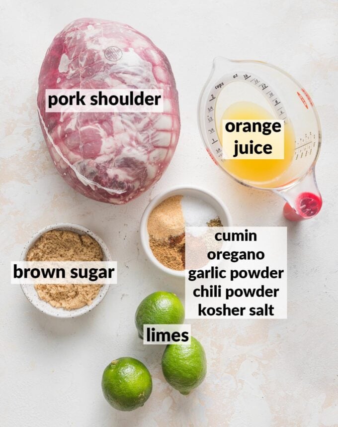 Labeled overhead photo of a boneless pork shoulder, still wrapped, orange juice, fresh limes, brown sugar, and spices, all in prep bowls and ready to cook.