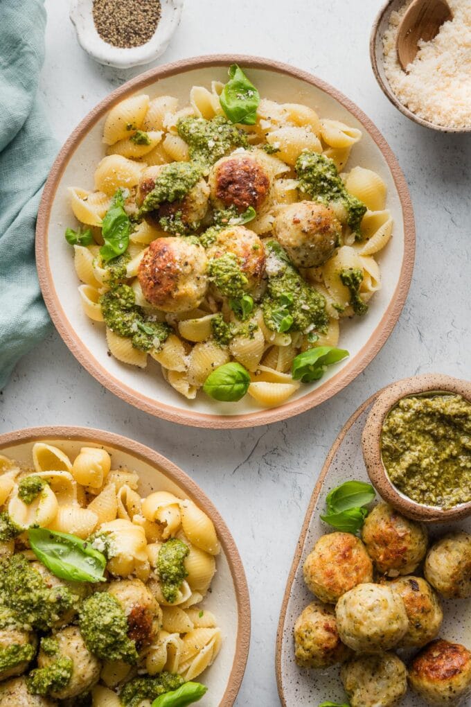 Plates of pasta with pesto chicken meatballs, fresh basil, extra pesto, and Parmesan.