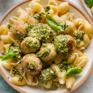 Bowl full of chicken pesto meatballs served with extra pesto, Parmesan, and basil leaves over medium shell pasta.