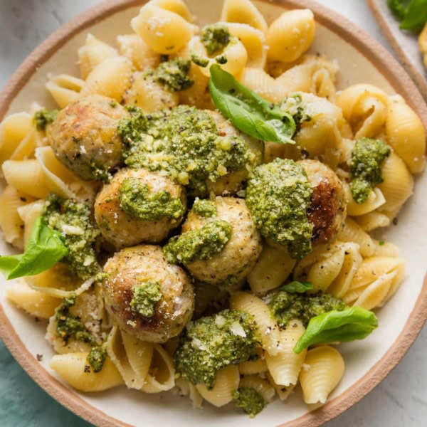 Bowl full of chicken pesto meatballs served with extra pesto, Parmesan, and basil leaves over medium shell pasta.