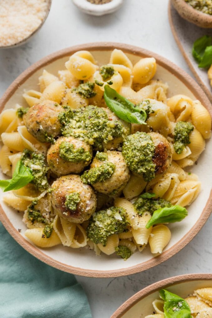Bowl full of chicken pesto meatballs served with extra pesto, Parmesan, and basil leaves over medium shell pasta.