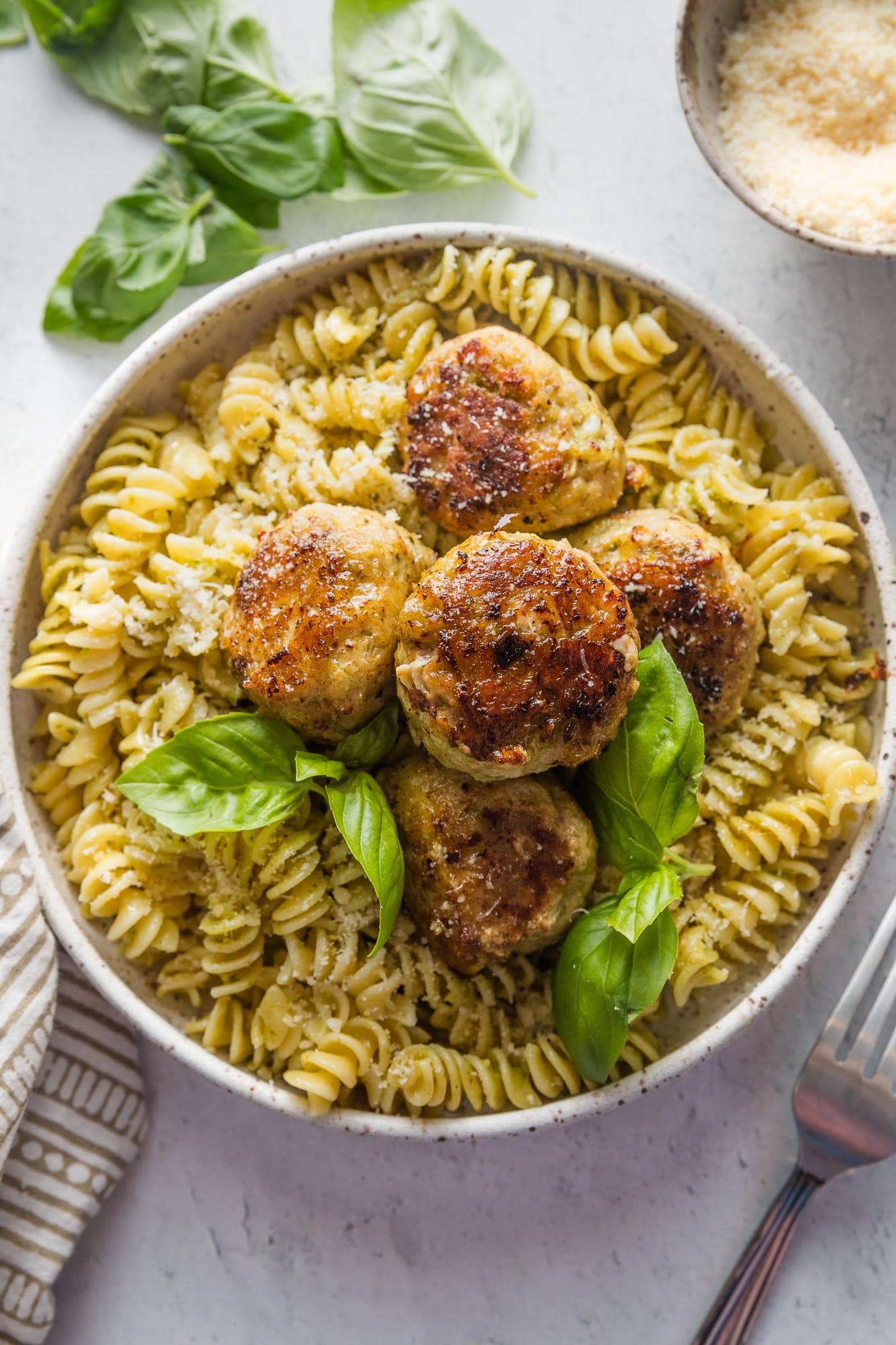 Chicken pesto meatballs served over rotini with Parmesan and fresh basil leaves.