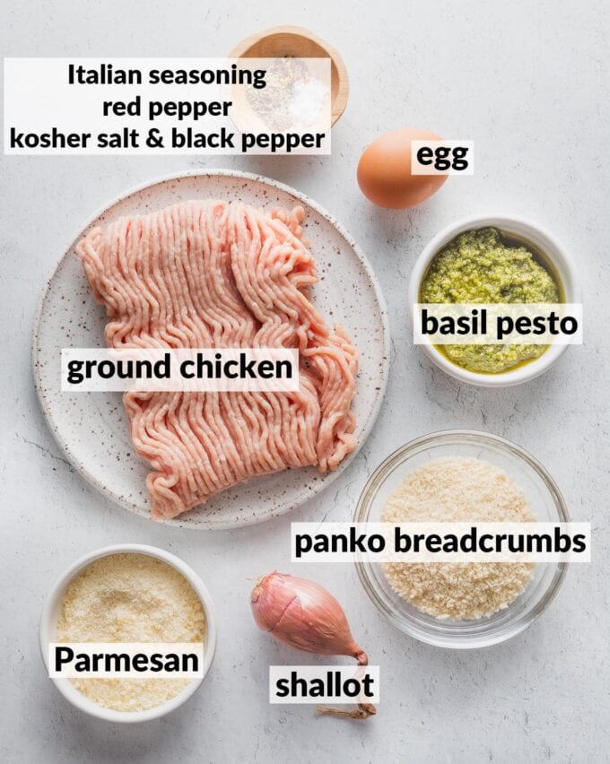 Labeled overhead image of ground chicken, basil pesto, panko breadcrumbs, Parmesan, shallot, egg, Italian seasoning, red pepper, kosher salt and black pepper, all in prep bowls and ready to mix into meatballs.
