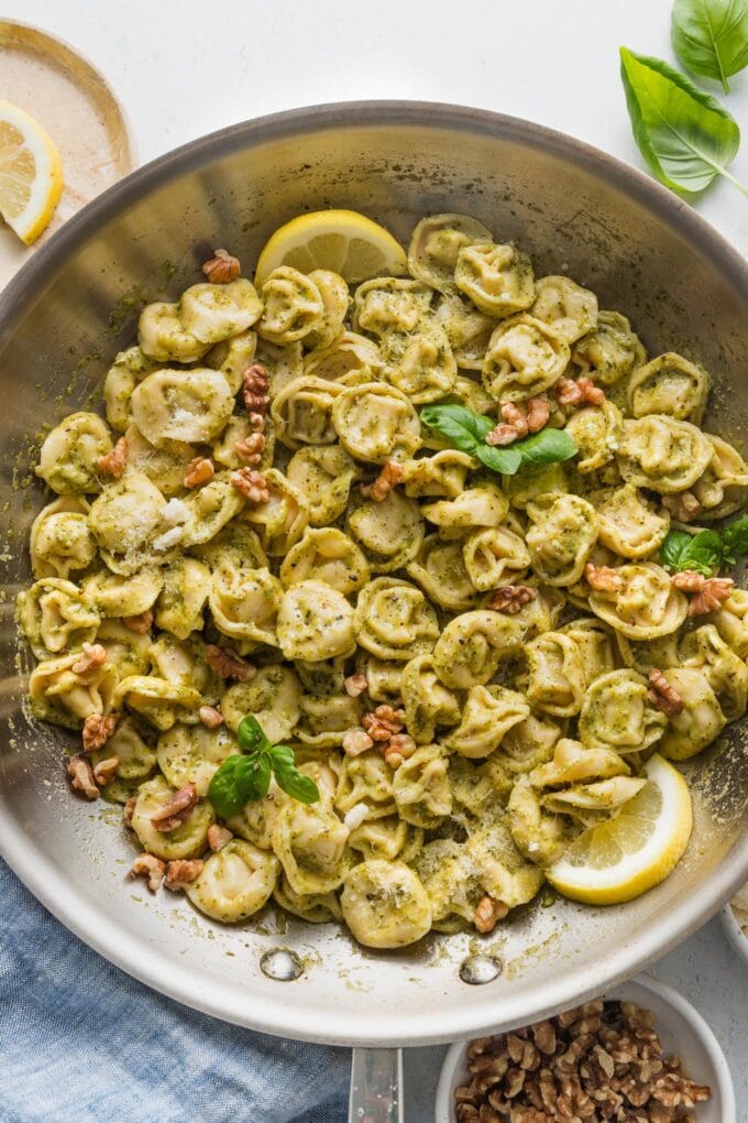Creamy pesto tortellini skillet with toasted walnuts, Parmesan cheese, lemon wedges, and fresh basil.