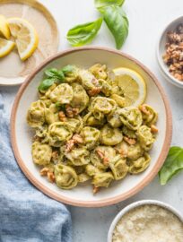 Low pasta bowl full of creamy pesto tortellini garnished with Parmesan and fresh basil leaves.