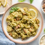 Countertop set with bowl of pesto tortellini and small plates with walnuts, cheese, lemon wedges, and fresh basil leaves.