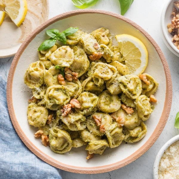 Countertop set with bowl of pesto tortellini and small plates with walnuts, cheese, lemon wedges, and fresh basil leaves.