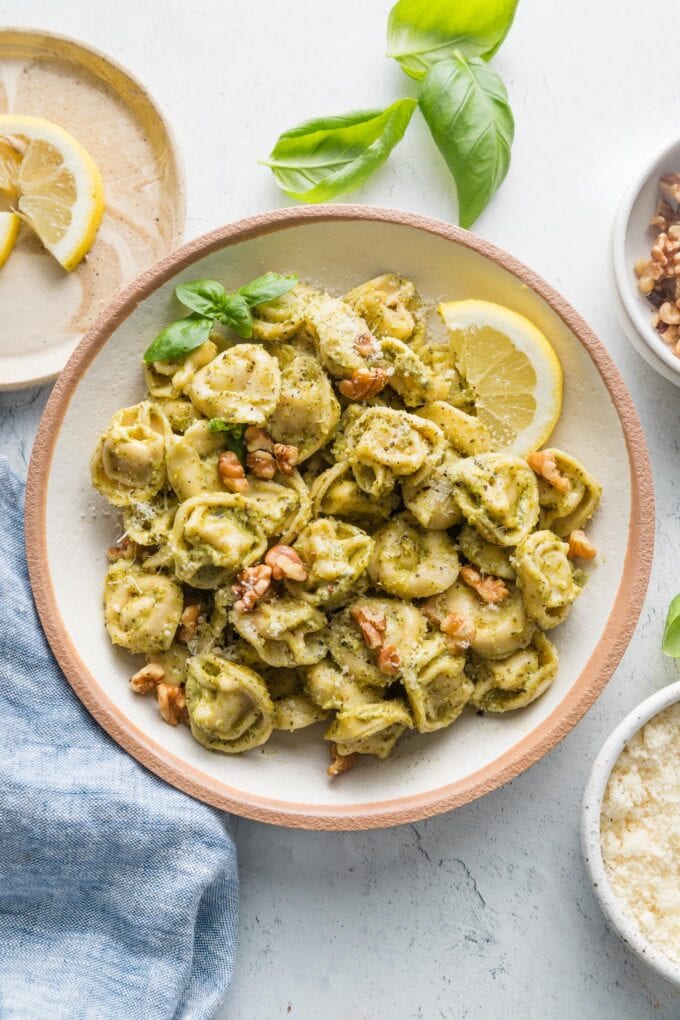 Countertop set with bowl of pesto tortellini and small plates with walnuts, cheese, lemon wedges, and fresh basil leaves.