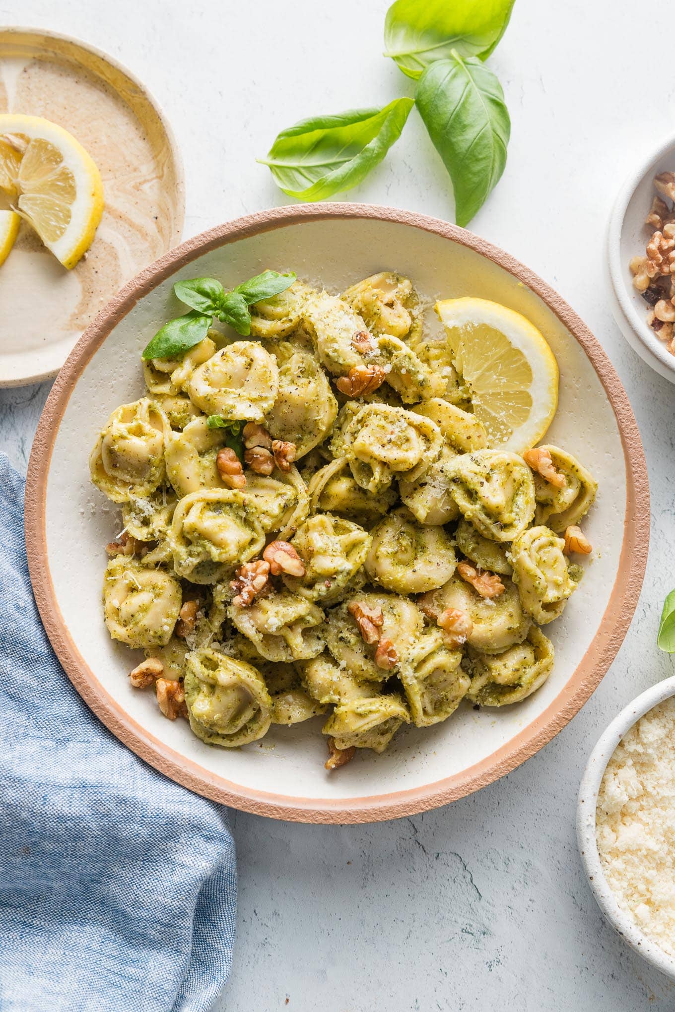 Countertop set with bowl of pesto tortellini and small plates with walnuts, cheese, lemon wedges, and fresh basil leaves.