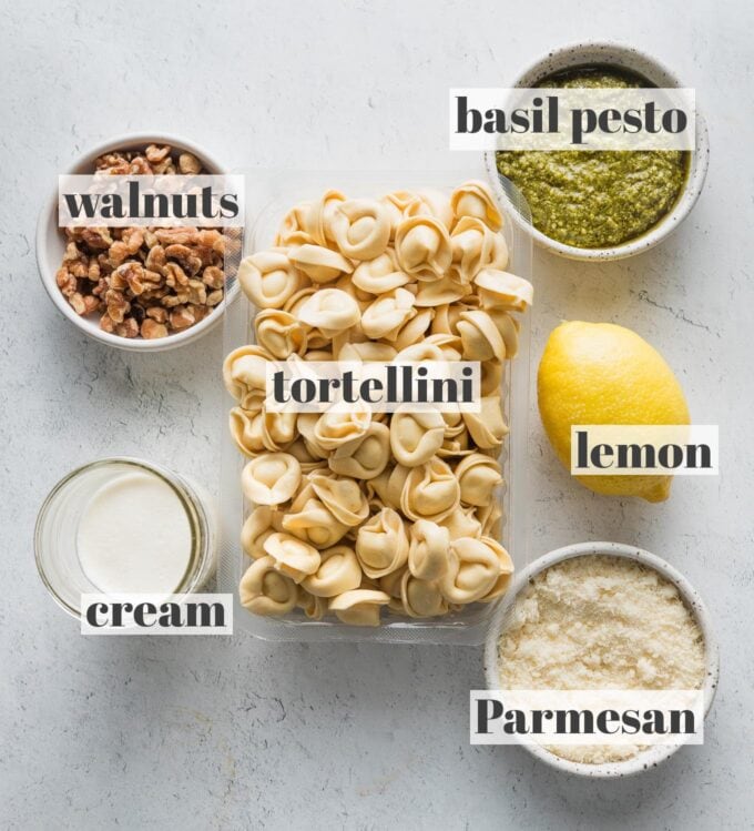 Labeled overhead image of refrigerated tortellini, basil pesto, chopped walnuts, a whole lemon, grated Parmesan cheese, and a small amount of heavy cream.