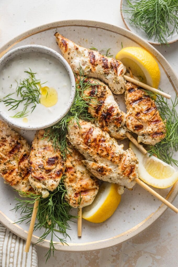 Serving platter piled high with Greek chicken skewers, lemon wedges, fresh dill, and a bowl of Tzatziki sauce for dipping.