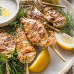 Close up of a large platter loaded with Greek chicken skewers, lemon wedges, Tzatziki sauce, and fresh dill.