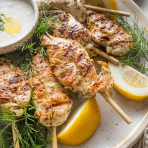 Close up of a large platter loaded with Greek chicken skewers, lemon wedges, Tzatziki sauce, and fresh dill.