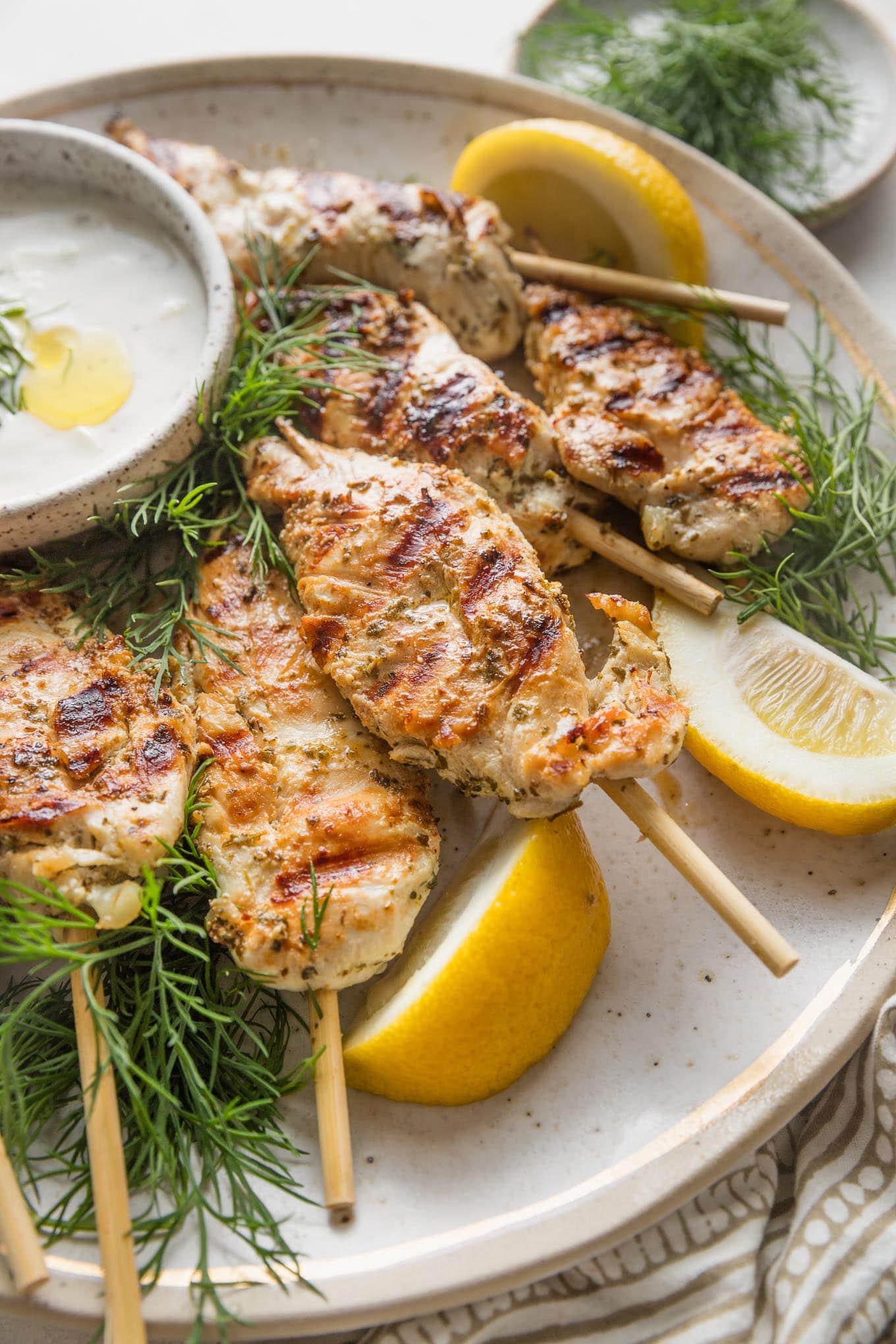 Close up of a large platter loaded with Greek chicken skewers, lemon wedges, Tzatziki sauce, and fresh dill.