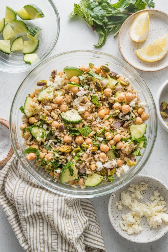 Clear glass bowl full of a salad with brown rice and chickpeas, surrounded by smaller bowls of chopped cucumber, crumbled feta, herbs, lemon wedges, and pistachios.