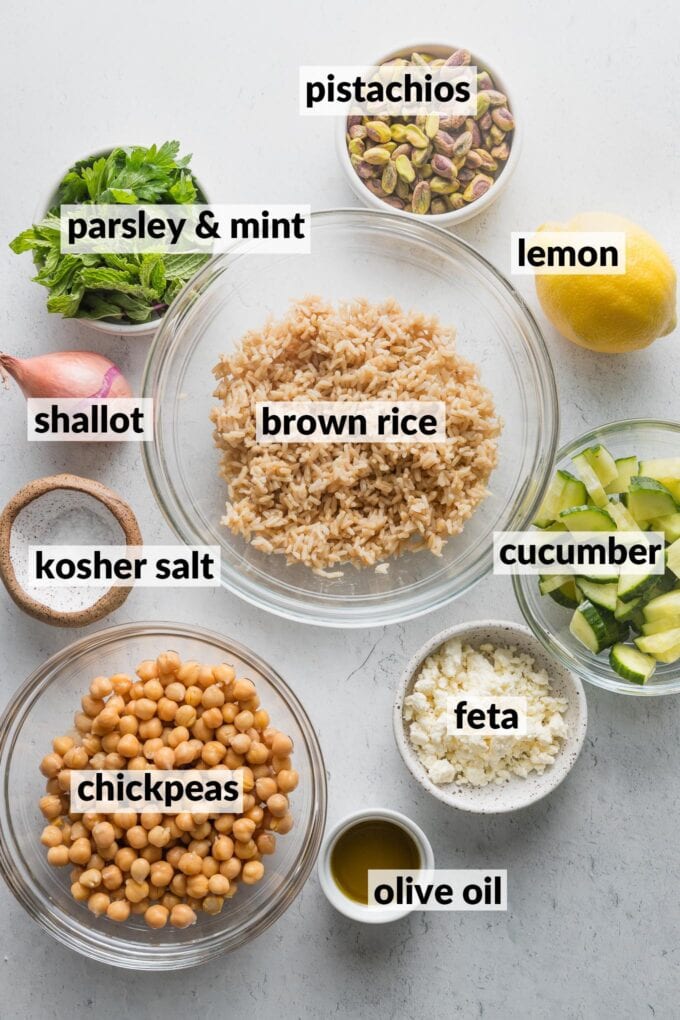 Labeled overhead photo of cooked brown rice, drained and rinsed chickpeas, fresh parsley and mint, pistachios, shallot, chopped cucumber, a whole lemon, olive oil, feta, and kosher salt, all in prep bowls and ready to combine into a salad.