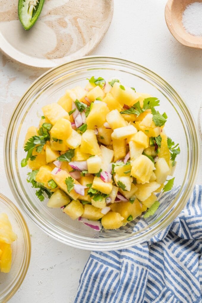 Small glass mixing bowl full of pineapple salsa, with extra pineapple, salt, and jalapeno on the side.