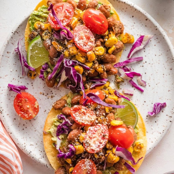Small plate holding two pinto bean tostadas topped with corn and tomato salsa, red cabbage, Cotija, and lime wedges.