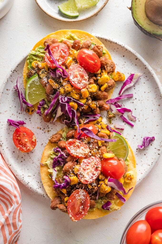 Small plate holding two pinto bean tostadas topped with corn and tomato salsa, red cabbage, Cotija, and lime wedges.