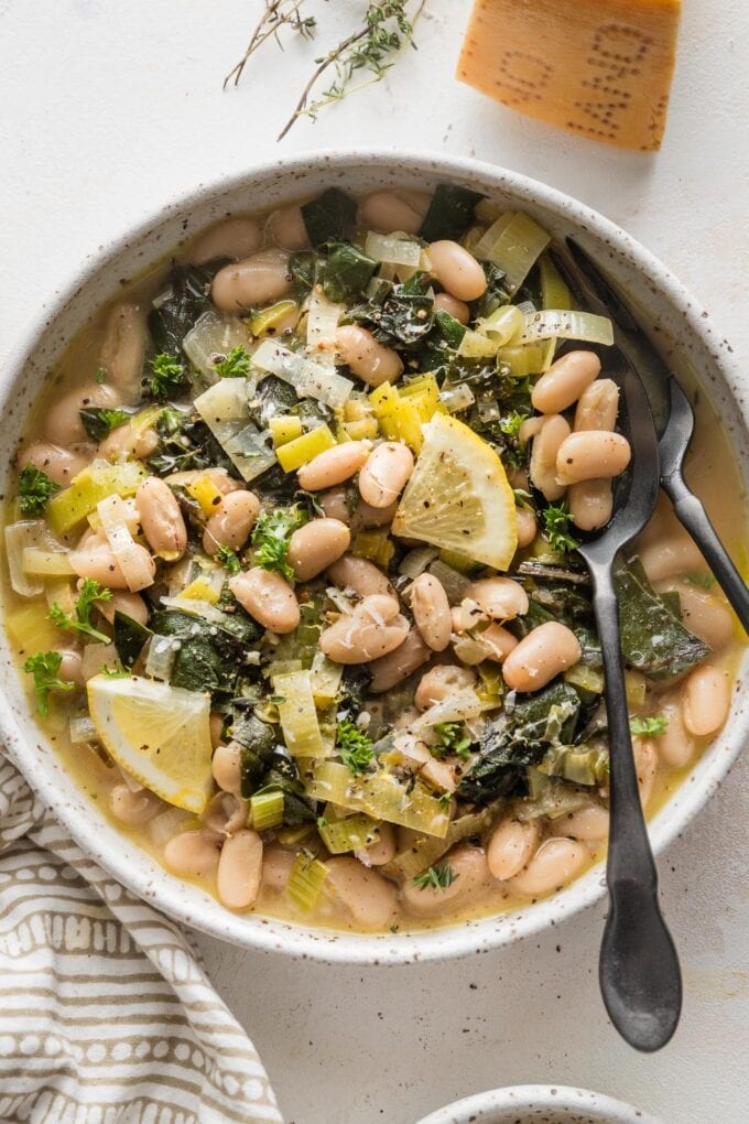 Small bowl of Mediterranean beans and greens with lemon wedges and a fork and spoon for serving.