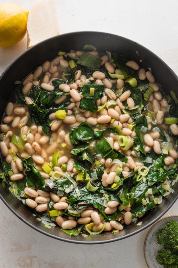 Large cast iron skillet filled with broth, beans, and greens, simmering slowly.