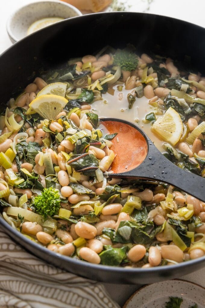 Spoon stirring a pot of Mediterranean beans and greens to show the cozy, light broth.