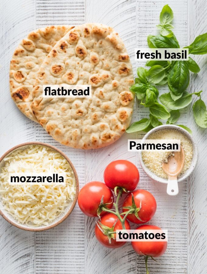 Labeled overhead view of naan, shredded mozzarella cheese, grated Parmesan, tomatoes on-the-vine, and fresh basil.
