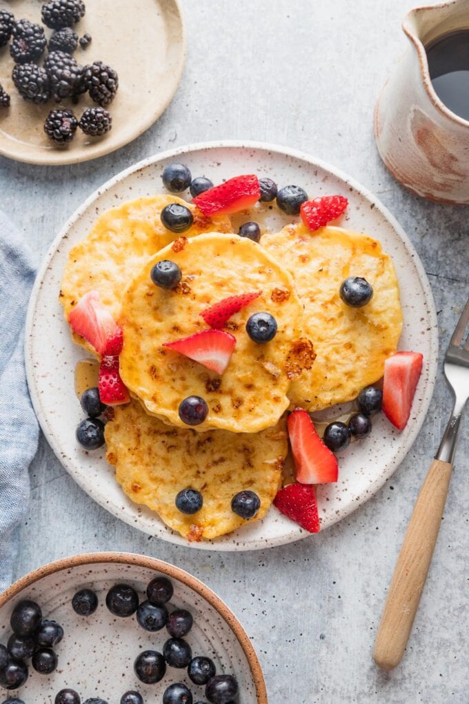 Countertop set with a plate of cottage cheese pancakes sprinkled with blueberries, strawberries, and maple syrup.