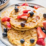 Plate piled with cottage cheese pancakes with fresh strawberries, blueberries, and a stream of maple syrup being drizzled on top.