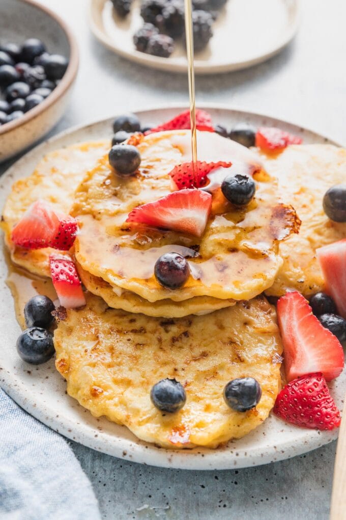 Plate piled with cottage cheese pancakes with fresh strawberries, blueberries, and a stream of maple syrup being drizzled on top.
