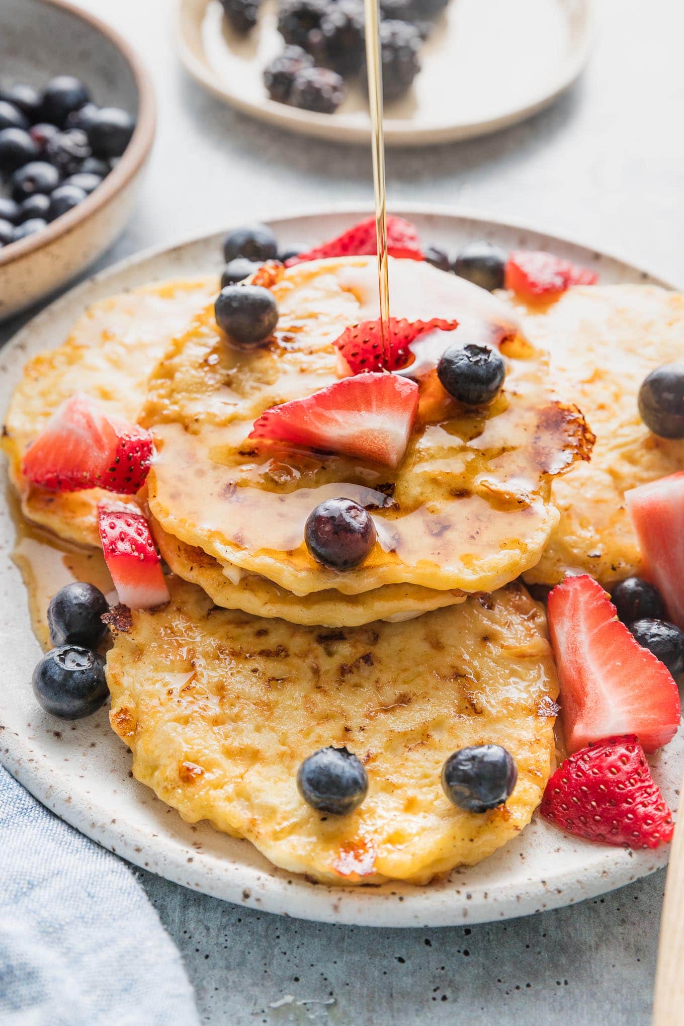 Plate piled with cottage cheese pancakes with fresh strawberries, blueberries, and a stream of maple syrup being drizzled on top.
