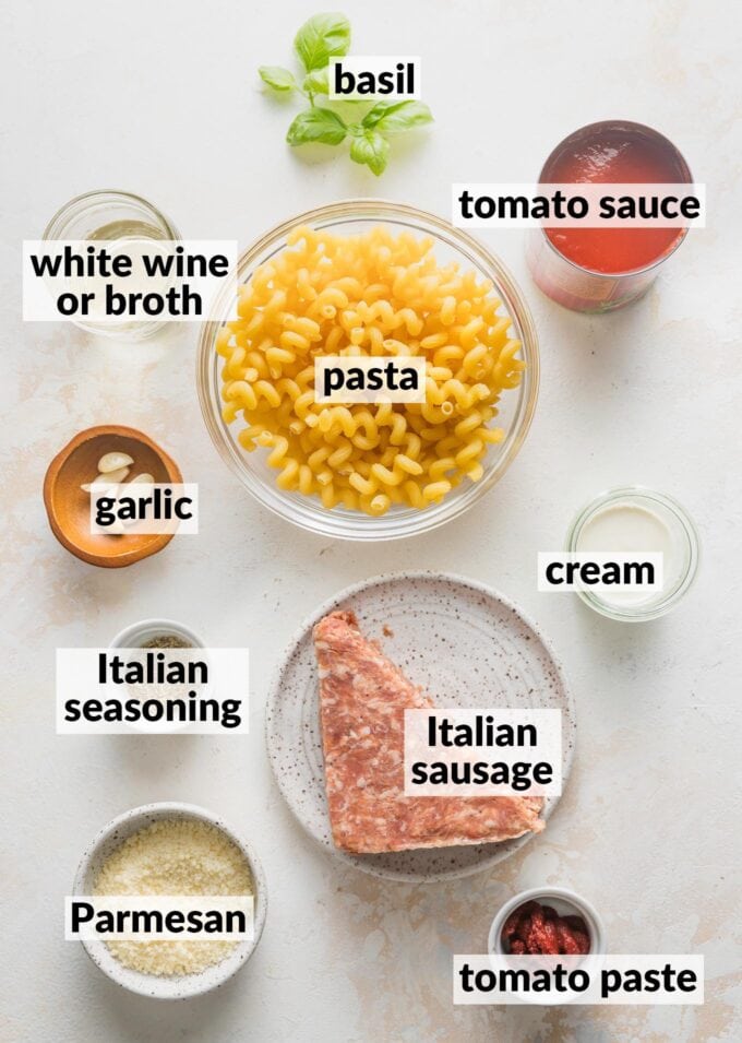 Labeled overhead photo of pasta, ground Italian sausage, tomato sauce, white wine, fresh basil, garlic cloves, cream, Italian seasoning, tomato pasta, and grated Parmesan cheese, all in prep bowls and ready to cook.
