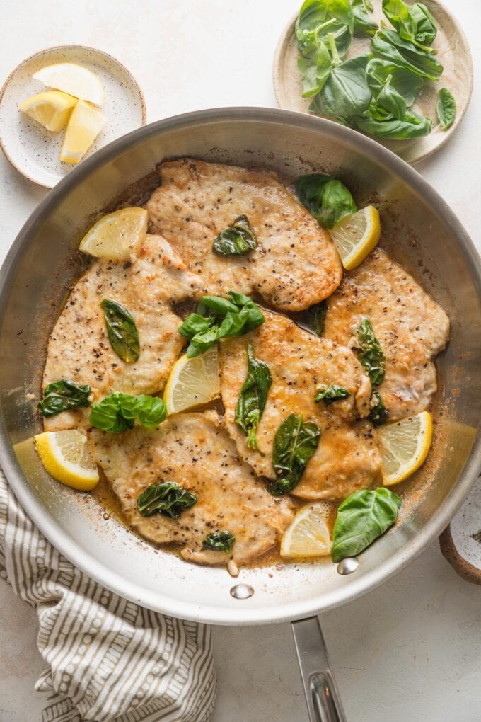 Skillet of lemon basil chicken with extra lemon wedges and basil scattered nearby.