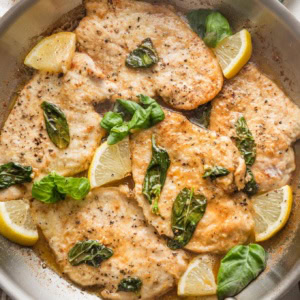 Large skillet full of lemon basil chicken breasts with extra lemon wedges on top as a garnish.