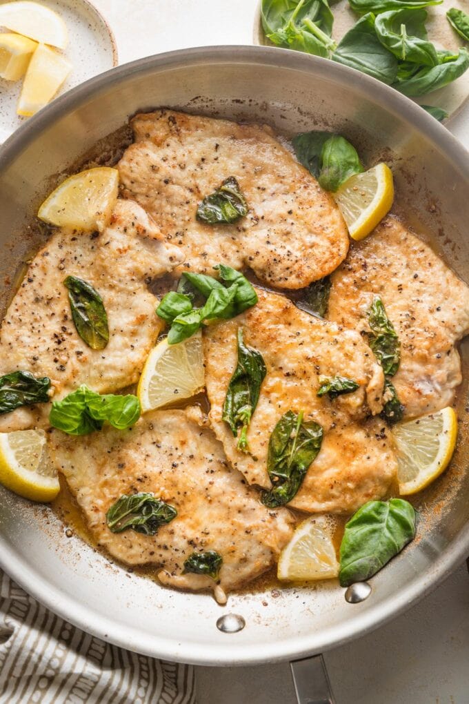 Large skillet full of lemon basil chicken breasts with extra lemon wedges on top as a garnish.