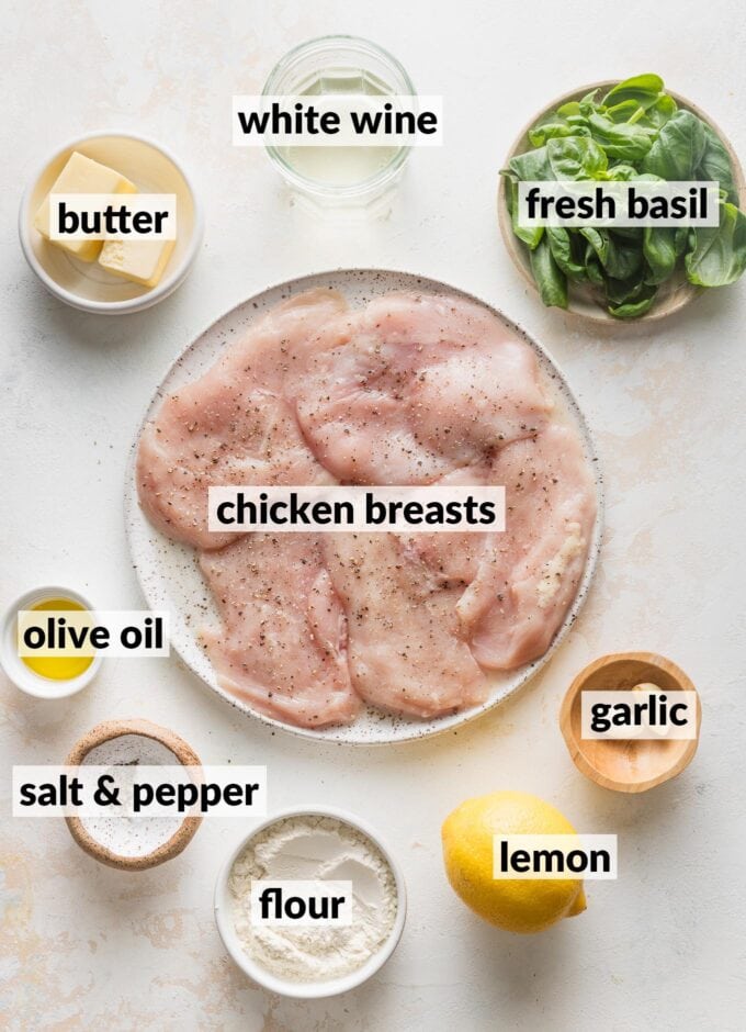 Labeled overhead image of chicken breasts seasoned with kosher salt and black pepper, a whole lemon, fresh basil, white wine, garlic, butter, and olive oil.