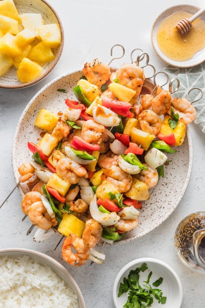Countertop set with a platter of shrimp pineapple skewers, extra pineapple chunks, honey, rice, and fresh parsley for garnish.