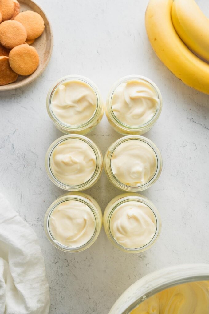 Six individual jars filled to the top with bananas, Nilla wafers, and a pudding-cream mixture, ready to go in the fridge.