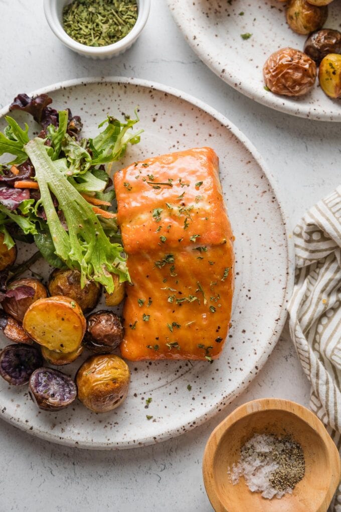 White ceramic plate filled with a fillet of brown sugar glazed salmon, salad greens, and roasted baby potatoes, with another plate, dried parsley, and extra salt and pepper in small bowls nearby on the table.
