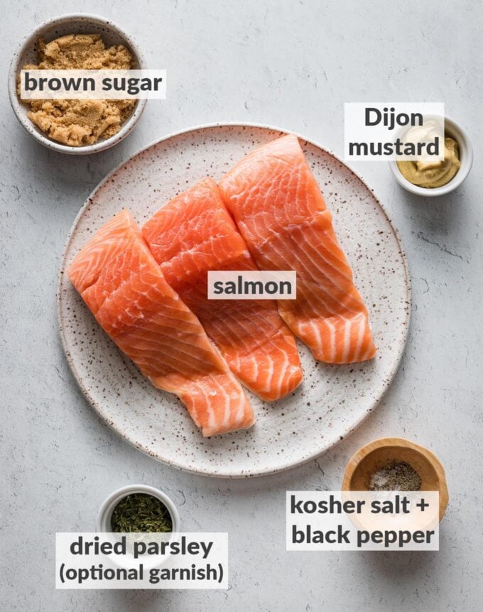 Labeled overhead photo of salmon fillets, brown sugar, Dijon mustard, kosher salt and black pepper, and dried parsley, all measured onto plates or prep bowls and ready to cook.