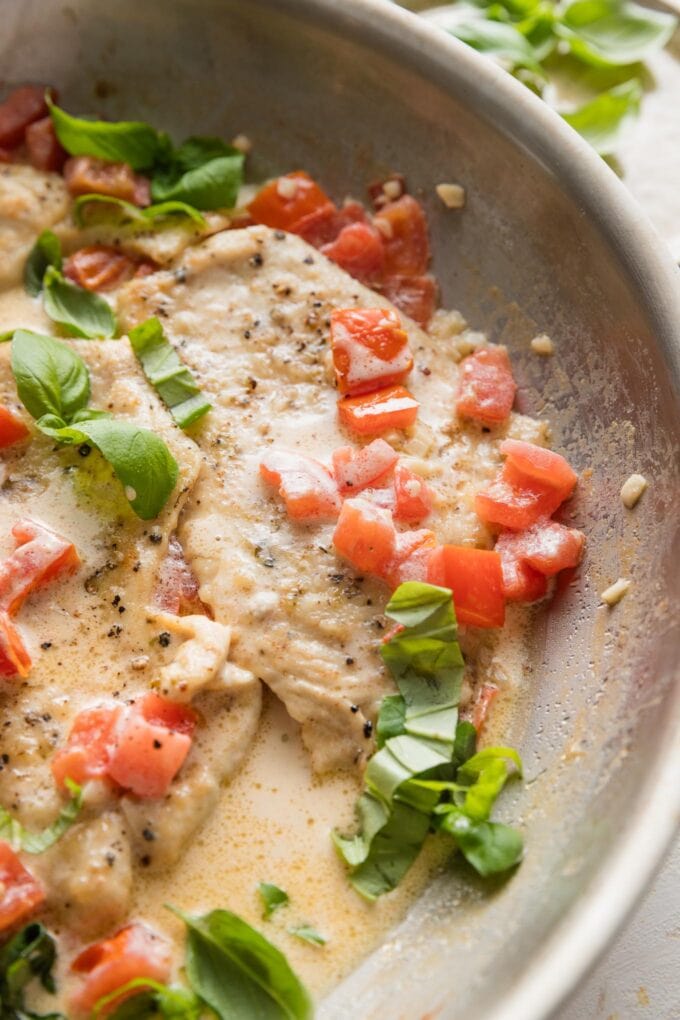 Close up of a thin-sliced chicken breast sauteed in a skillet with a light cream sauce, fresh diced tomatoes, and fresh basil.