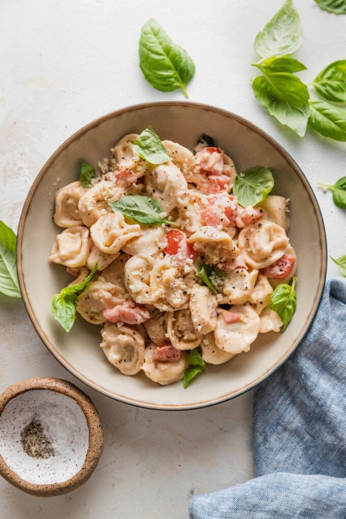 Bowl full of creamy tomato basil tortellini with extra salt, pepper, and basil leaves scattered around.