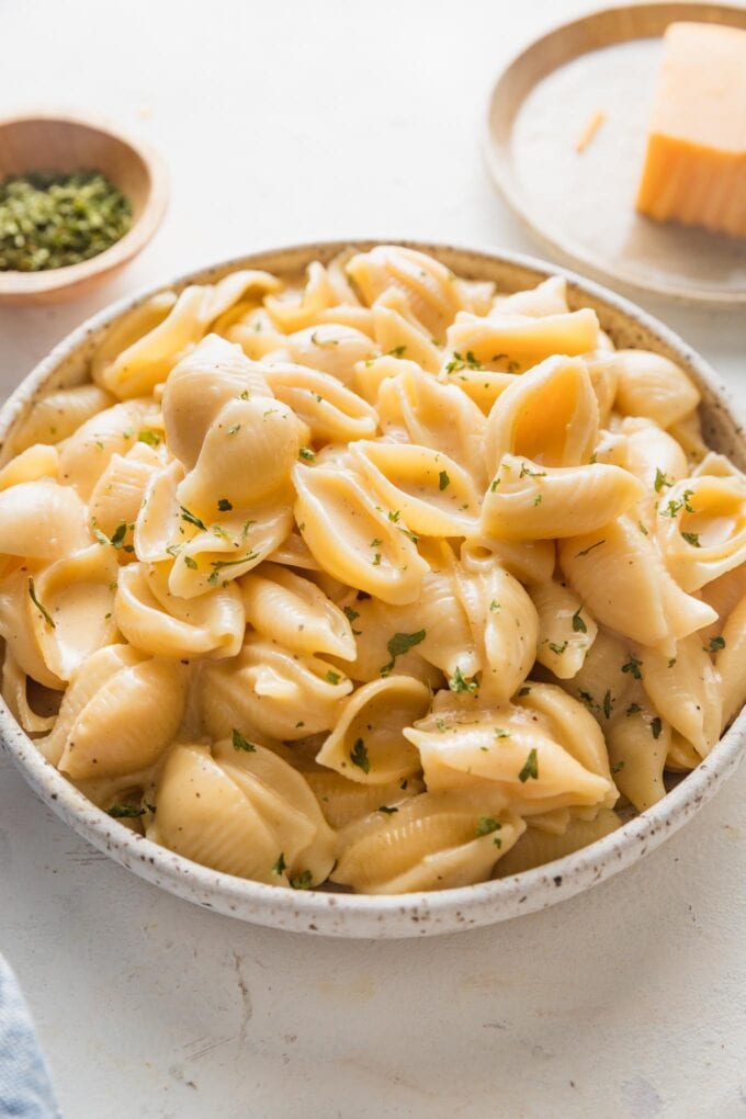 Angled view of a countertop set with a pasta bowl full of mac and cheese with extra cheddar cheese and dried parsley in the background.