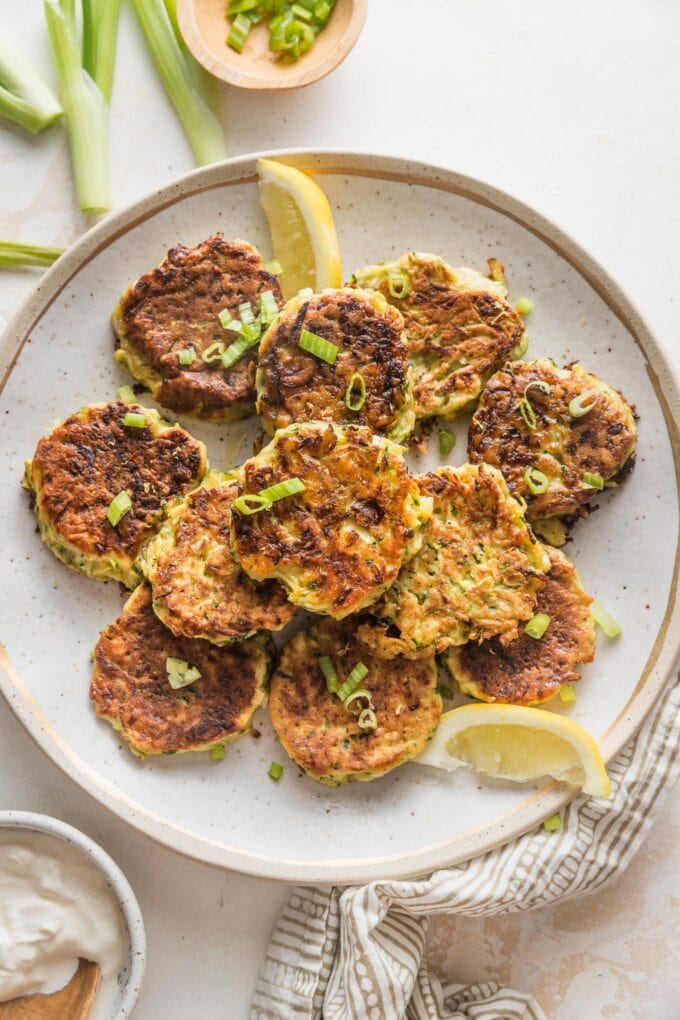 Large plate filled with zucchini fritters garnished with sliced green onions and lemon wedges.